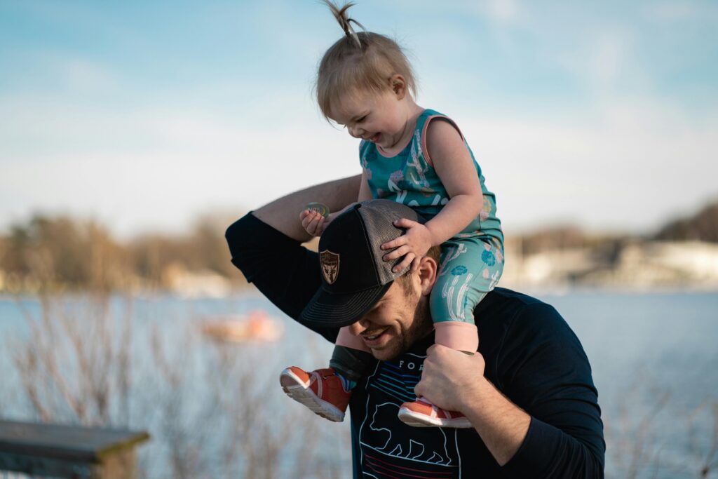 Father carrying daughter on shoulders by the lake, sharing a joyful moment together.