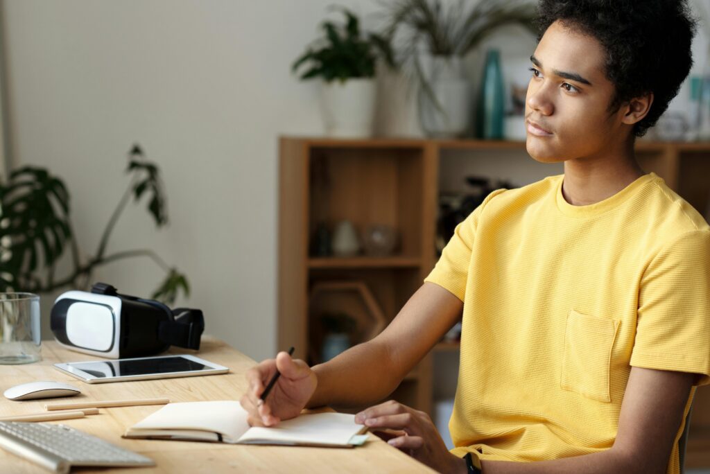 Teenage boy in a yellow shirt studying indoors, focused and pensive, with a notebook and tablet on a wooden table.