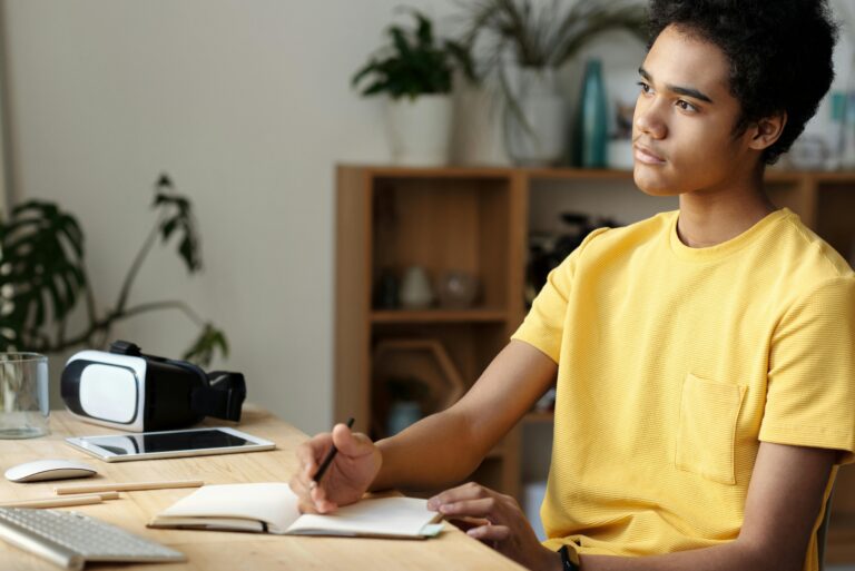 Teenage boy in a yellow shirt studying indoors, focused and pensive, with a notebook and tablet on a wooden table.